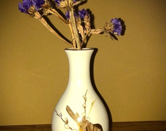 Puede incluir: Un jarrón de cerámica blanco con un pájaro dorado y un diseño floral, que contiene un ramo de flores secas moradas.