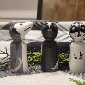 Puede incluir: Tres figuras de animales de madera pintadas a mano: una zarigüeya, un zorrillo y un mapache. La zarigüeya es gris y blanca con una nariz rosa. El zorrillo es blanco y negro. El mapache es blanco y negro con una cara pintada.