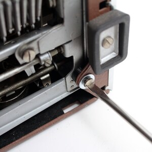 May include: A close-up of a vintage typewriter with a screwdriver being used to tighten a screw on the side of the machine. The typewriter is brown and silver with a black rubber foot.