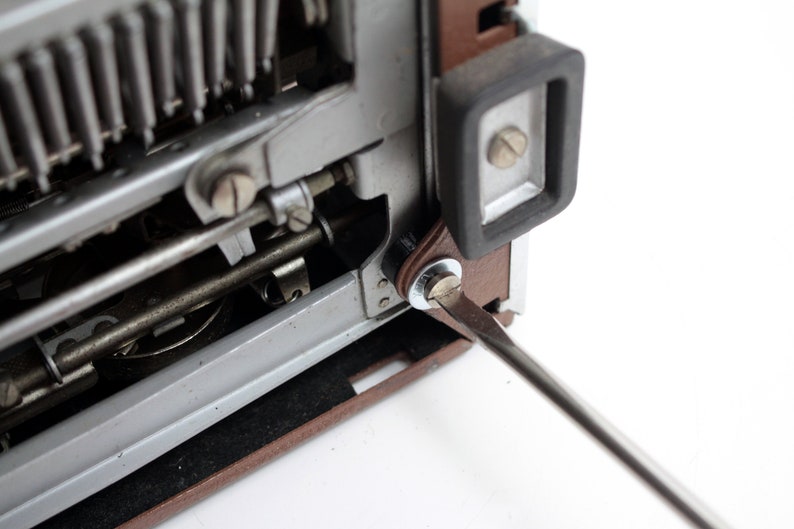May include: A close-up of a vintage typewriter with a screwdriver being used to tighten a screw on the side of the machine. The typewriter is brown and silver with a black rubber foot.