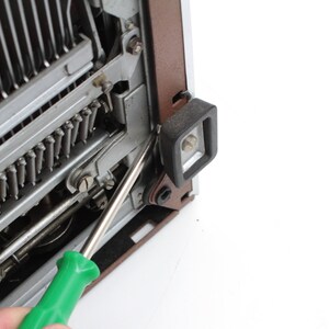 May include: A green screwdriver is being used to remove a screw from a typewriter. The screw is located on the side of the typewriter, near the carriage return lever.