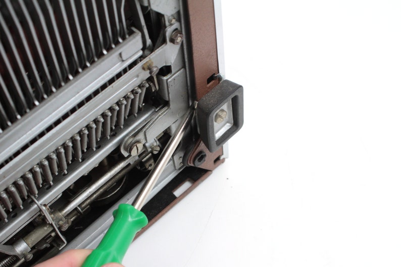 May include: A green screwdriver is being used to remove a screw from a typewriter. The screw is located on the side of the typewriter, near the carriage return lever.