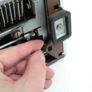 May include: Close-up of a typewriter mechanism with a hand holding a small metal part. The typewriter is brown and silver with a black rubber foot.