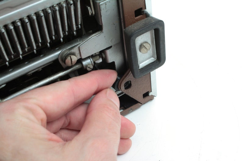 May include: Close-up of a typewriter mechanism with a hand holding a small metal part. The typewriter is brown and silver with a black rubber foot.
