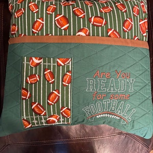 May include: Green and brown quilted pillow with the text "Are you ready for some football" embroidered on it. The pillow has a pocket on the front with a football field design and footballs.