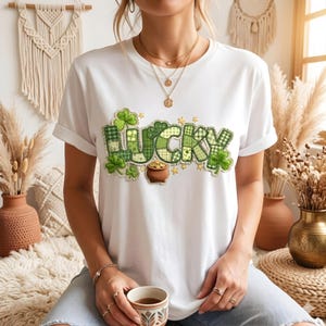 May include: White t-shirt featuring the word "LUCKY" in green patchwork lettering, shamrocks, and a pot of gold. The design includes gold stars, ideal for St. Patrick's Day celebrations.