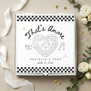 Könnte beinhalten: Weiße Pizzabox mit einem schwarz-weißen Schachbrettmuster-Rand. Die Box zeigt den Text "That's Amore" über einer herzförmigen Pizza-Illustration. Darunter stehen die Namen "Danielle & Todd" und das Datum "11. Juli 2026".