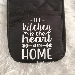 May include: Black quilted oven mitt with white text that reads "The kitchen is the heart of the home".