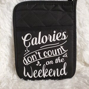 May include: Black quilted oven mitt with a pocket and a hanging loop. The mitt features the white text "Calories don't count on the Weekend" in a whimsical font. The background is a fluffy white surface.