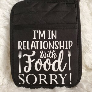 May include: Black quilted oven mitt with white text that reads "I'm in a relationship with food, sorry!" with a fork graphic on either side of the word "food".