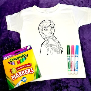 Op de afbeelding: Een wit T-shirt met een zwart-witte lijntekening van een meisje met lange vlechten. Het shirt is ontworpen om in te kleuren met stiften. Een pak van 10 Crayola-stiften is inbegrepen. De stiften zitten in een gele en oranje doos met de tekst "Crayola Markers" en "Voorkeur van leraren!" op de voorkant.