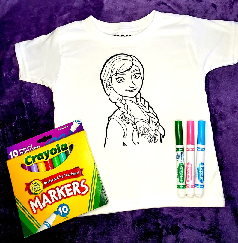Op de afbeelding: Een wit T-shirt met een zwart-witte lijntekening van een meisje met lange vlechten. Het shirt is ontworpen om in te kleuren met stiften. Een pak van 10 Crayola-stiften is inbegrepen. De stiften zitten in een gele en oranje doos met de tekst "Crayola Markers" en "Voorkeur van leraren!" op de voorkant.