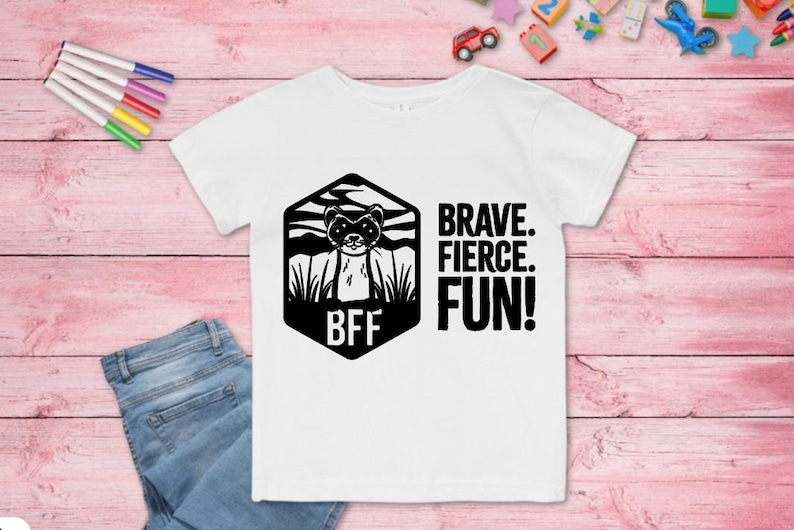 Puede incluir: Camiseta blanca con un gr&aacute;fico en blanco y negro de un hur&oacute;n dentro de un hex&aacute;gono con las letras "BFF". Las palabras "BRAVE. FIERCE. FUN!" est&aacute;n impresas en negro. Tambi&eacute;n se ven unos vaqueros azules y rotuladores de colores.