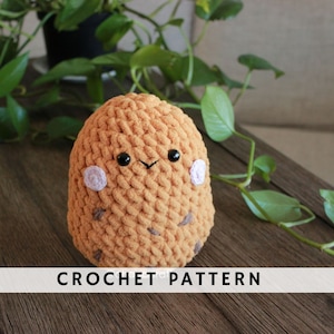 Peut inclure: Un jouet en peluche en forme de pomme de terre crocheté avec un corps brun, des yeux noirs et des joues roses. Le jouet est posé sur une surface en bois.
