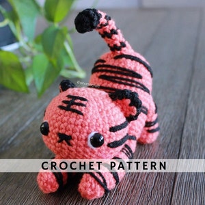 May include: Crochet pattern for a pink and black tiger amigurumi. The tiger is lying on its belly with its tail curled up. The pattern is available for purchase.