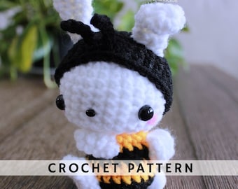 May include: A crocheted bee amigurumi toy with black and yellow stripes, white body, and black antennae. The toy has large black eyes and pink cheeks. The toy is sitting on a wooden surface. CROCHET PATTERN is written in white text on a black background.