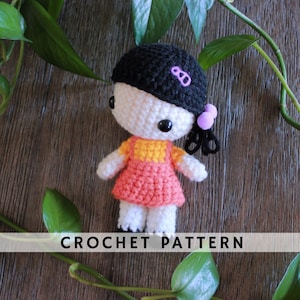 May include: Crochet pattern for a small doll with black hair, a pink dress, and a yellow top. The doll has large eyes and a sweet expression. The pattern is available for download.