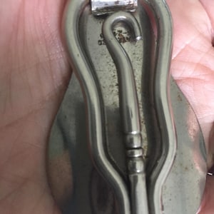 May include: A vintage silver-toned money clip with a curved design. The clip features a hook-shaped element and a decorative central bar. The clip is held in a hand, showing the back of the clip.