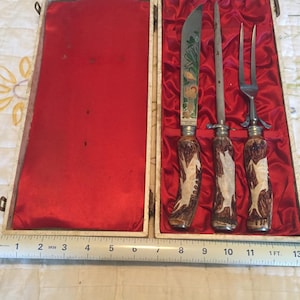 May include: An antique carving set with a knife, a sharpening steel, and a fork, presented in a cream-colored box with a red satin lining. The handles feature a deer design. The knife blade has a decorative etched design.