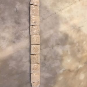 May include: Ivory-colored bracelet composed of rectangular, carved beads with various embossed designs. The bracelet is connected with a metal clasp and a small chain. The beads feature symbols and patterns, suggesting a cultural or historical significance.