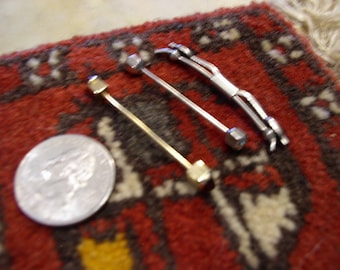 May include: Three metal tie pins in gold and silver tones, with hexagonal and skeletal designs, are displayed on a red and brown patterned rug. A silver coin is also present, adding scale to the image.
