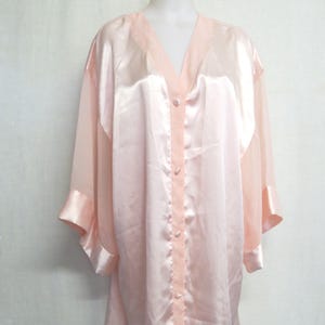 Puede incluir: Una bata de satén rosa claro con cuello en V y cierre de botones. La bata tiene mangas largas con paneles transparentes y puños a juego. La prenda se exhibe en un maniquí blanco.