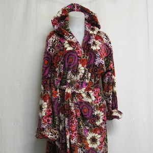 Puede incluir: Bata con capucha con estampado floral en tonos rojos, rosas, morados y blancos. La bata tiene mangas largas y cinturón anudado. El diseño floral se repite. Se muestra en un maniquí.