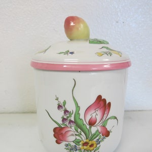 May include: White ceramic jar with a pink lid and floral design. The lid has a small, green leaf and a pink and yellow fruit on top. The jar has a floral design with pink, yellow, and green flowers.