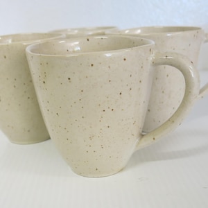 Può includere: Un set di quattro tazze in ceramica maculata. Le tazze sono di colore beige chiaro con piccole macchie marroni. Ogni tazza ha un manico arrotondato e un bordo leggermente svasato. Le tazze sono disposte in gruppo.