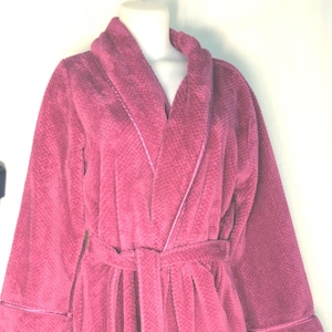 May include: A plush, magenta-colored robe with a shawl collar and a matching belt. The robe has long sleeves with a contrasting trim around the cuffs and collar. The fabric appears soft and textured, ideal for lounging.