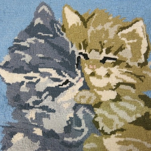 May include: A close-up of a needlepoint pillow featuring two kittens, one gray and one brown, with their heads touching. The background is a light blue color.