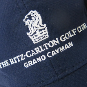 May include: A navy blue baseball cap featuring white embroidered text that reads THE RITZ-CARLTON GOLF CLUB GRAND CAYMAN, accompanied by a white lion crest logo. A white Under Armour logo is visible on the side panel of the cap.