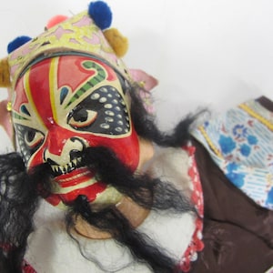 May include: A traditional Chinese puppet with a colorful painted face mask, black hair, and a red, white, and blue patterned costume.