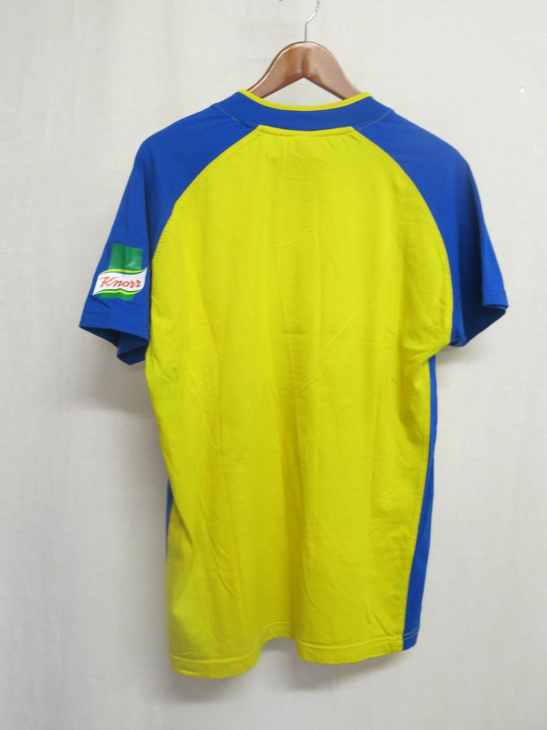 May include: Yellow and blue short-sleeved t-shirt with a green and red Knorr logo on the left sleeve.
