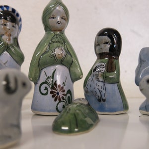 May include: A nativity scene set with hand-painted ceramic figurines. The set includes Mary, Joseph, baby Jesus, a sheep, and two lambs. The figurines are painted in shades of blue, green, and white.