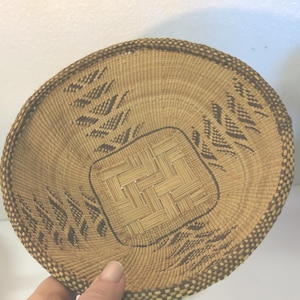 May include: A woven basket with a round shape and a flat base. The basket is made of natural-colored fibers with a decorative pattern of dark brown leaf-like shapes around the edges and a square woven design in the center.