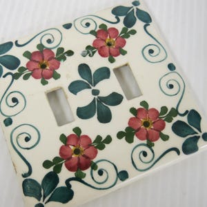 Pode incluir: Uma placa de interruptor duplo de cerâmica em tom esbranquiçado, de formato quadrado, com um padrão pintado à mão de flores cor-de-rosa com centros amarelos, folhas verdes e detalhes de vinhas em espiral num tom azul-petróleo escuro.