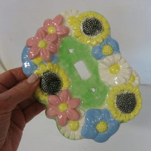 May include: A ceramic light switch cover decorated with colorful flowers. The cover features a green center with a switch opening, surrounded by pink, yellow, white, and blue flowers. The flowers have detailed petals and dark centers.