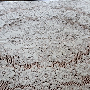 May include: Cream-colored lace tablecloth with a floral pattern. The intricate design features repeating floral and geometric shapes, creating a delicate and elegant texture. The lace is laid over a brown surface, enhancing the details.
