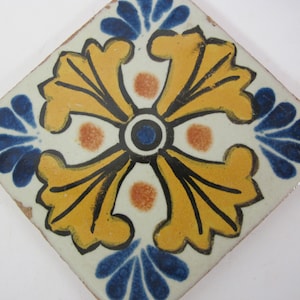 May include: A square ceramic tile featuring a yellow, blue and white floral design. The tile has a central motif of a four-pointed star with a blue and white centre. The star is surrounded by four yellow floral shapes with blue accents.