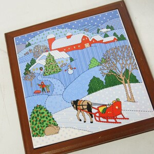 May include: A framed tile featuring a winter scene with a red barn, snow-covered hills, and a horse-drawn sleigh. The artwork includes a snowman, evergreen trees, and a rabbit. The frame is made of dark wood.