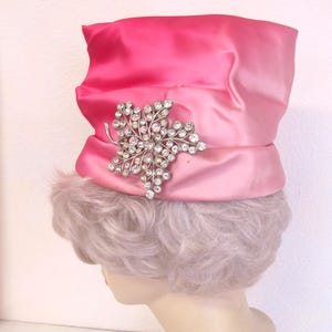 Op de afbeelding: Een roze satijnen hoed met een gradi&euml;nteffect, versierd met een sprankelende zilverkleurige bladbroche. De hoed heeft een brede rand en een decoratieve band. De hoed wordt getoond op een mannequinhoofd.