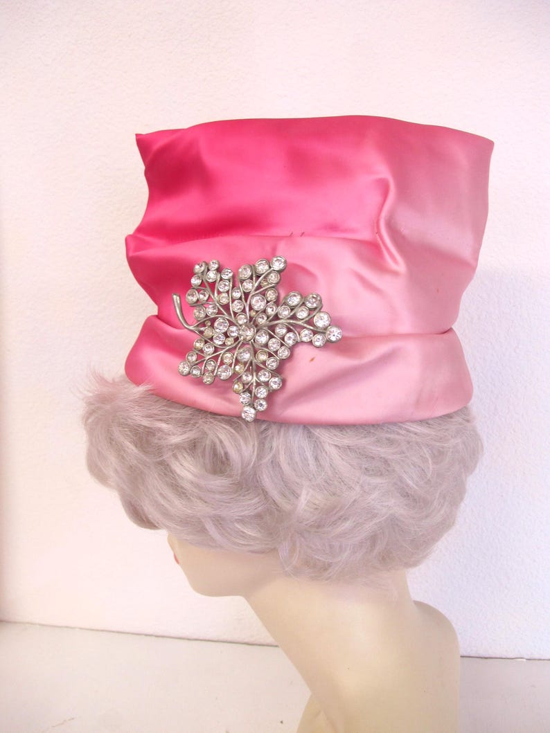 Op de afbeelding: Een roze satijnen hoed met een gradi&euml;nteffect, versierd met een sprankelende zilverkleurige bladbroche. De hoed heeft een brede rand en een decoratieve band. De hoed wordt getoond op een mannequinhoofd.