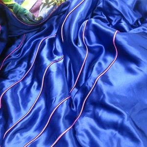 May include: Blue satin fabric with pink piping. The fabric is folded and draped, showing the texture and sheen of the satin.