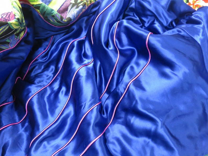 May include: Blue satin fabric with pink piping. The fabric is folded and draped, showing the texture and sheen of the satin.