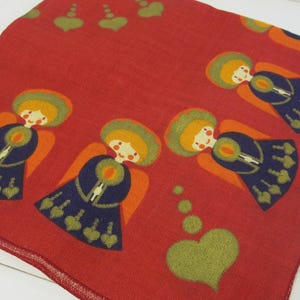 Può includere: Tessuto rosso con un motivo ripetuto di angeli con capelli gialli, vestiti arancioni e blu e ali verdi. Lo sfondo ha un motivo di cuori e cerchi verdi.