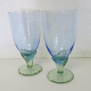 May include: Two blue-tinted glass goblets with a textured surface. The stems are a combination of green and blue, with a flower-like design at the base of the bowl. The foot of the goblets is a light green color.