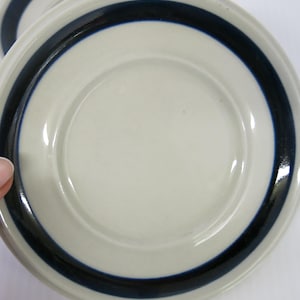 May include: Two off-white ceramic plates with a dark blue band around the rim. The plates have a simple, minimalist design. The plates are round and appear to be in good condition. The plates are likely for serving food.