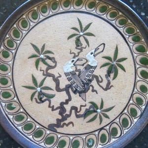 Op de afbeelding: Een ronde keramische plaat met een bruine rand en een beige midden. De plaat heeft een decoratieve rand met groene en witte accenten. Het middenontwerp omvat twee vogels, groene bladeren en bruine takken.