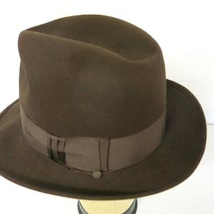 Puede incluir: Un sombrero fedora marrón con una banda marrón oscuro y un pequeño detalle de botón. El sombrero tiene una forma clásica y estructurada y un ala ancha. Este accesorio es elegante.